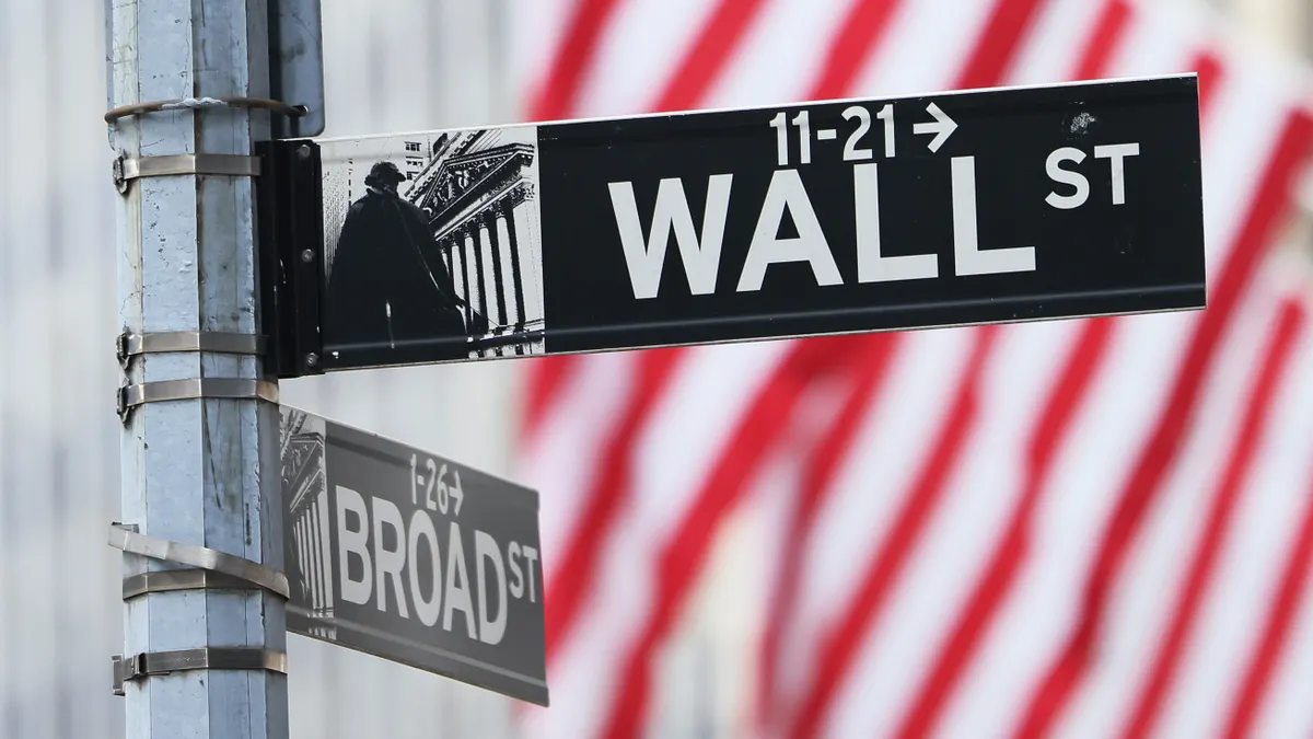 StreetsignsfortheintersectionofWallSt.andBroadSt.,withthestripesofanAmericanflaginthebackground.