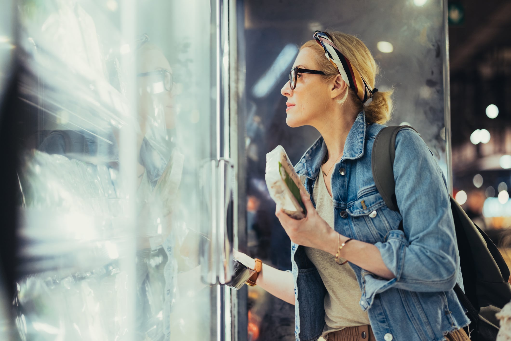 Close up shot of a blonde woman with blonde hair tied in a bun, wearing black glasses. She opened a fridge and is looking inside choosing products to buy.